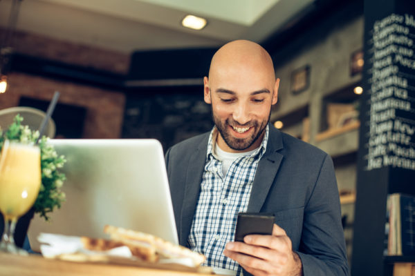 Un homme à une table de restaurant, regarde son téléphone portable et travaille sur son ordinateur portable